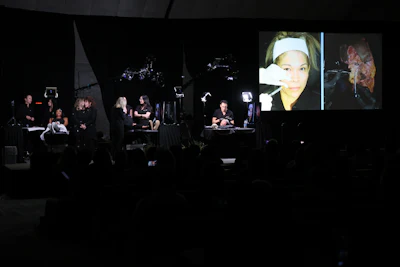 Several dermatologists are assessing the cosmetic needs of several patients on a stage and the video appears on a big screen.