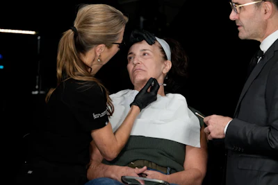 Two dermatologists are injecting botox into an older woman's jowl area.