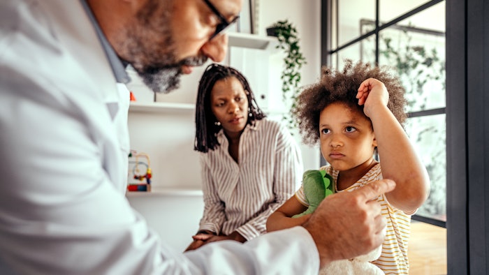 This is a photo of a pediatrician or dermatologist checking a rash on the arm of a Black toddler as his mother watches in worry.