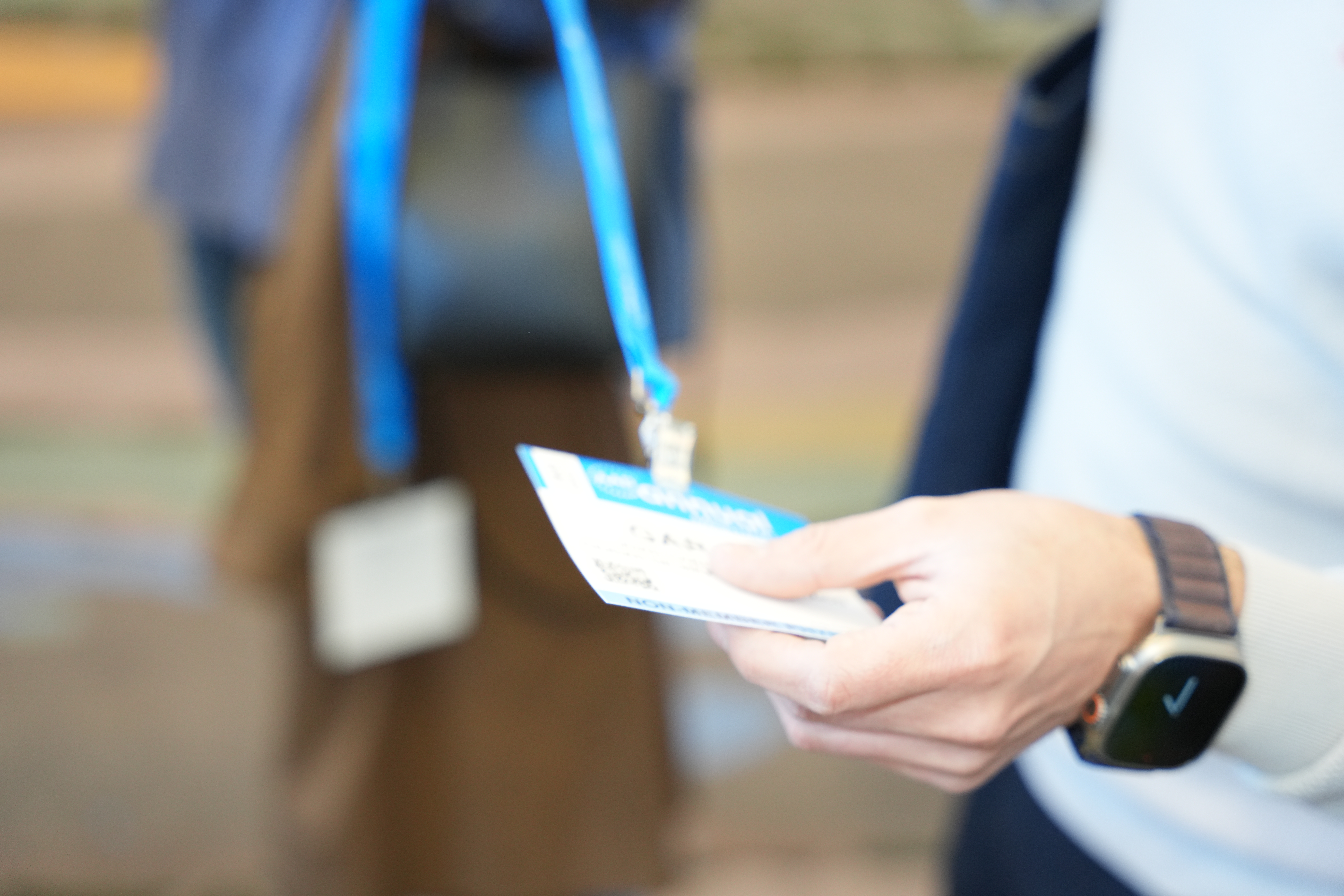 Conference badge with blue lanyard held by hand wearing watch