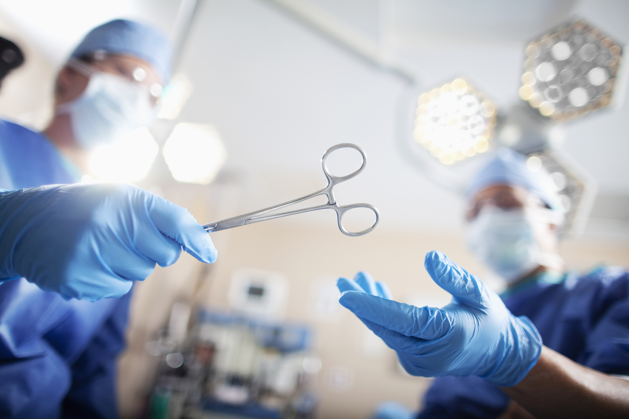 Surgeon in blue gloves holding surgical scissors in operating room with overhead lights and medical equipment