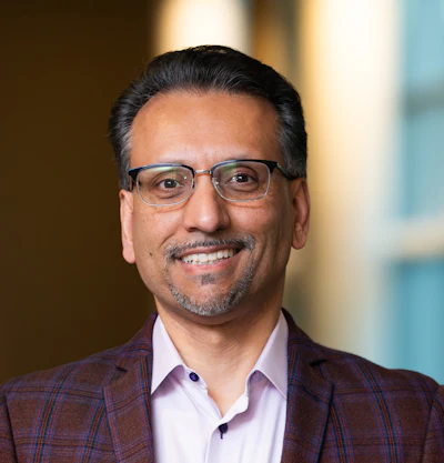Professional headshot of a man wearing glasses and burgundy plaid blazer with light pink shirt, smiling against blurred office background