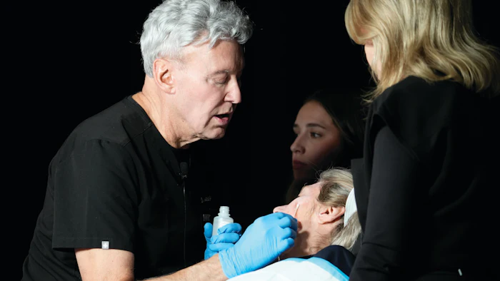 Medical professional in blue gloves demonstrating a cosmetic or skincare procedure to two women in a clinical setting
