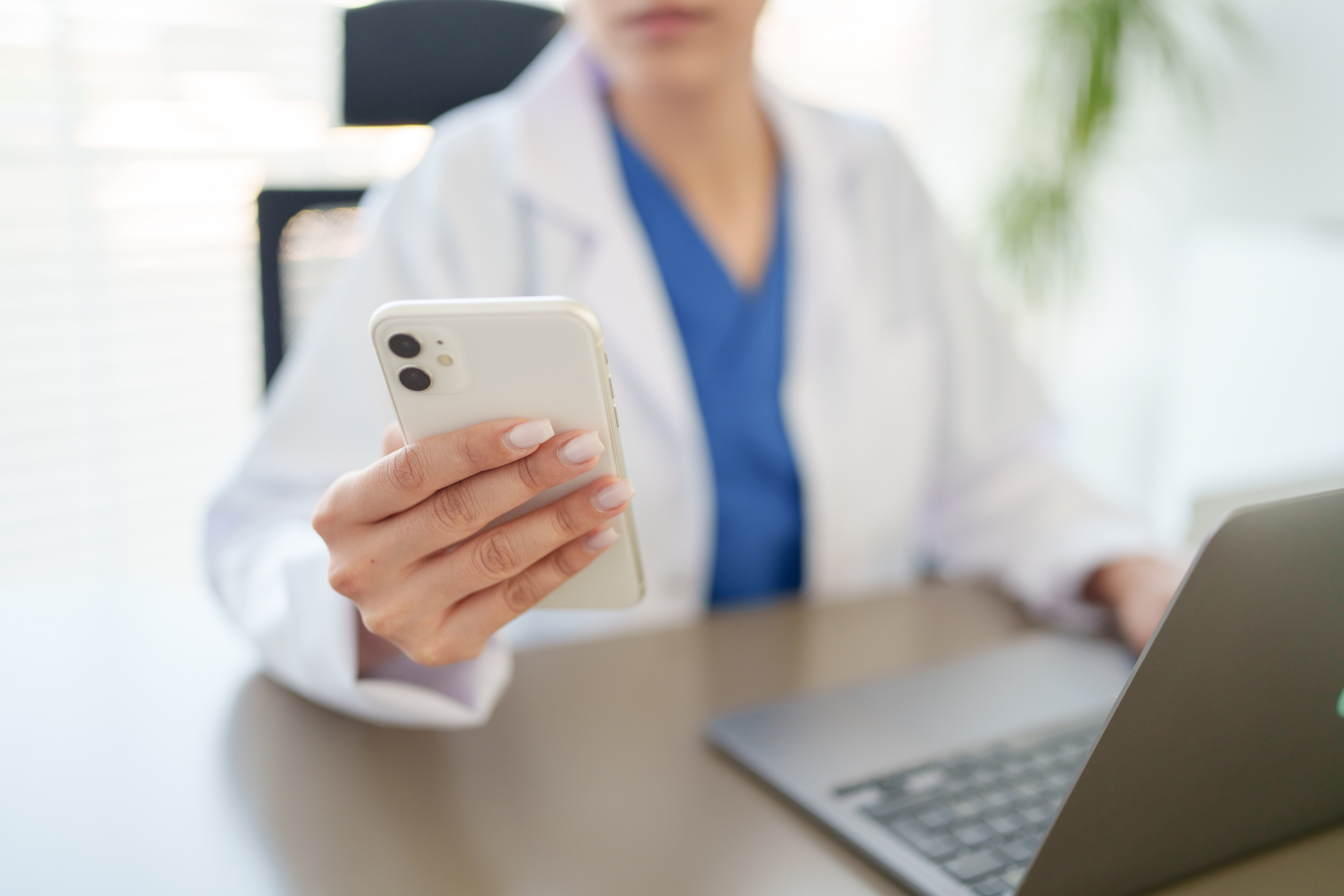 Healthcare professional holding white iPhone in medical office with laptop and plants