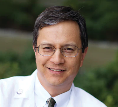 Professional headshot of a man wearing glasses and white shirt with tie, smiling at camera with green blurred background