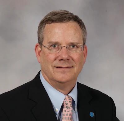 Professional headshot of a man in glasses wearing a black blazer, light blue shirt, and patterned tie