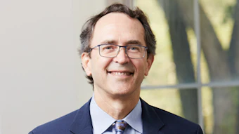 Professional headshot of a man wearing glasses and dark blazer with blue tie