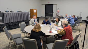 Professional conference attendees seated at round tables during a panel discussion or networking event in a meeting room.
