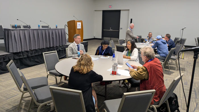 Professional conference attendees seated at round tables during a panel discussion or networking event in a meeting room.