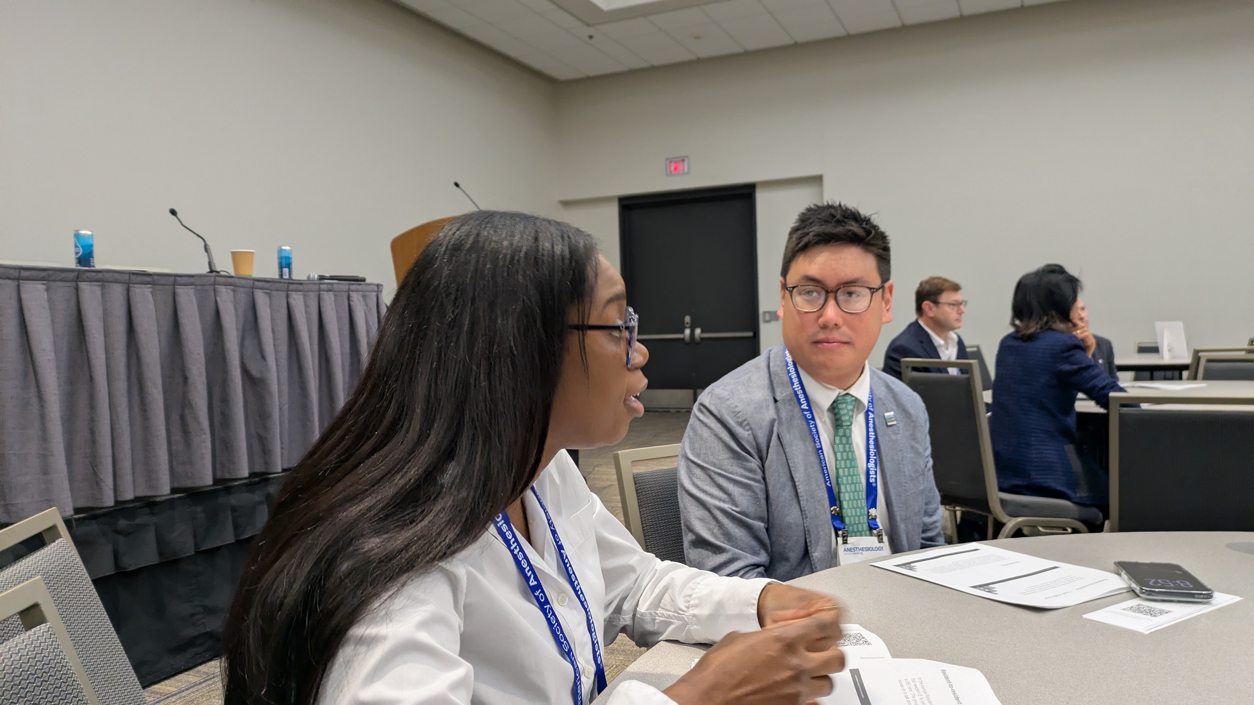 Two professionals wearing conference badges sit at a table during a business meeting or networking event