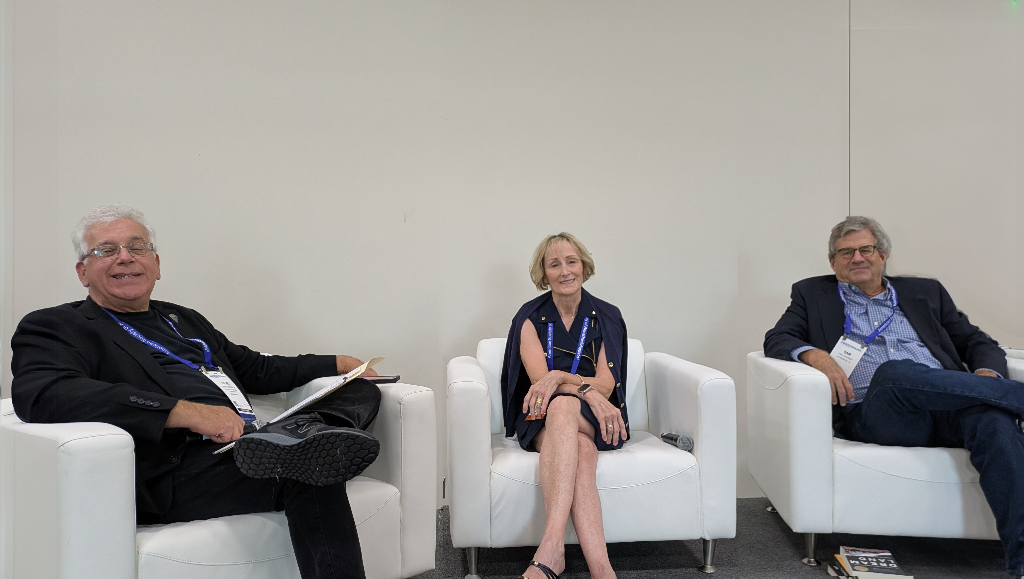 Three professionals seated on white chairs at a conference panel discussion event