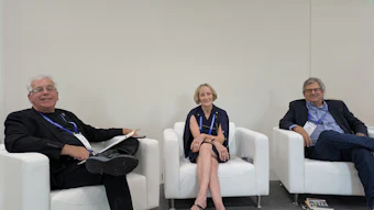 Three professionals seated on white chairs at a conference panel discussion event
