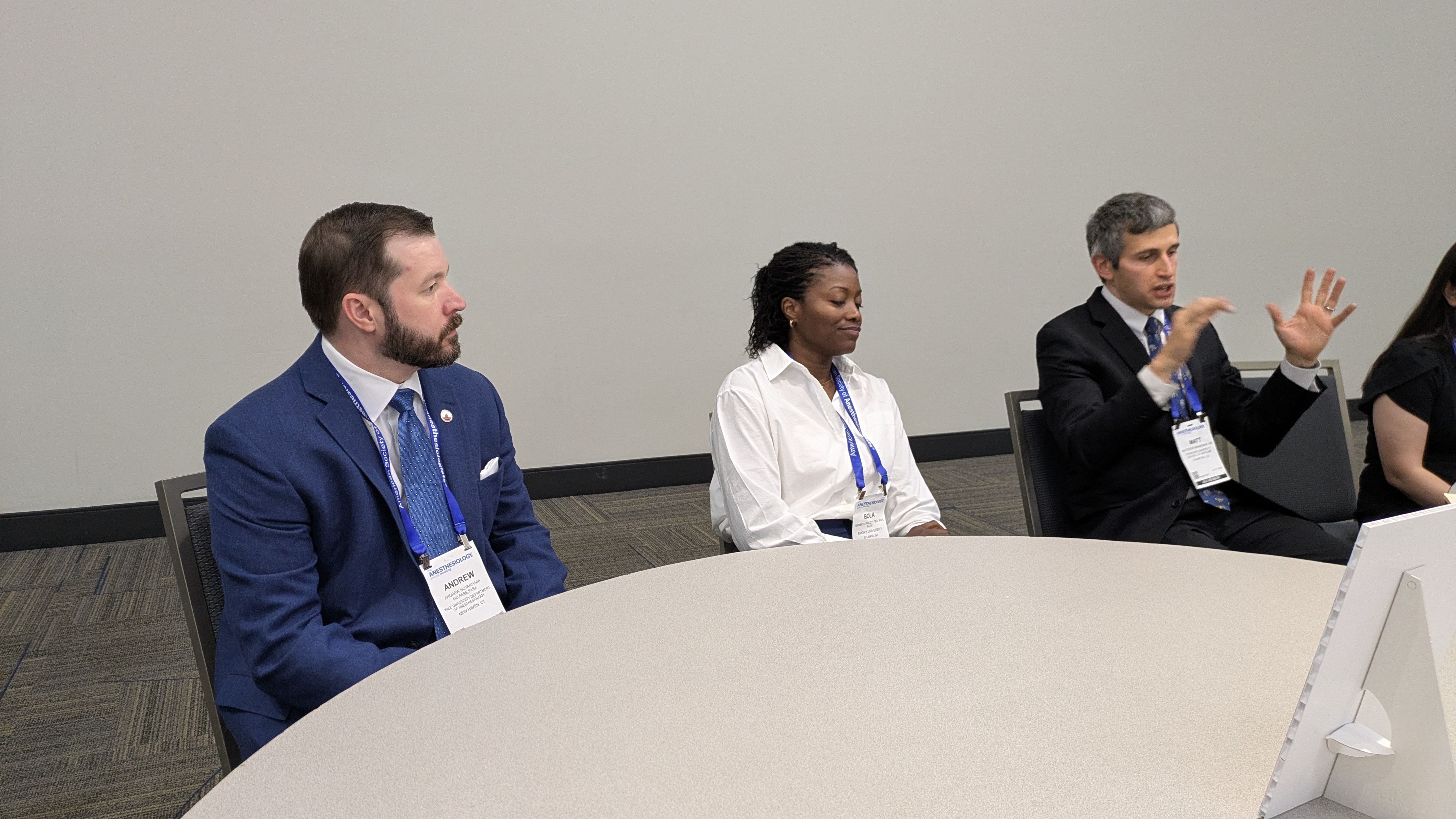Three professionals in business attire participate in a panel discussion at a conference table with credential badges.
