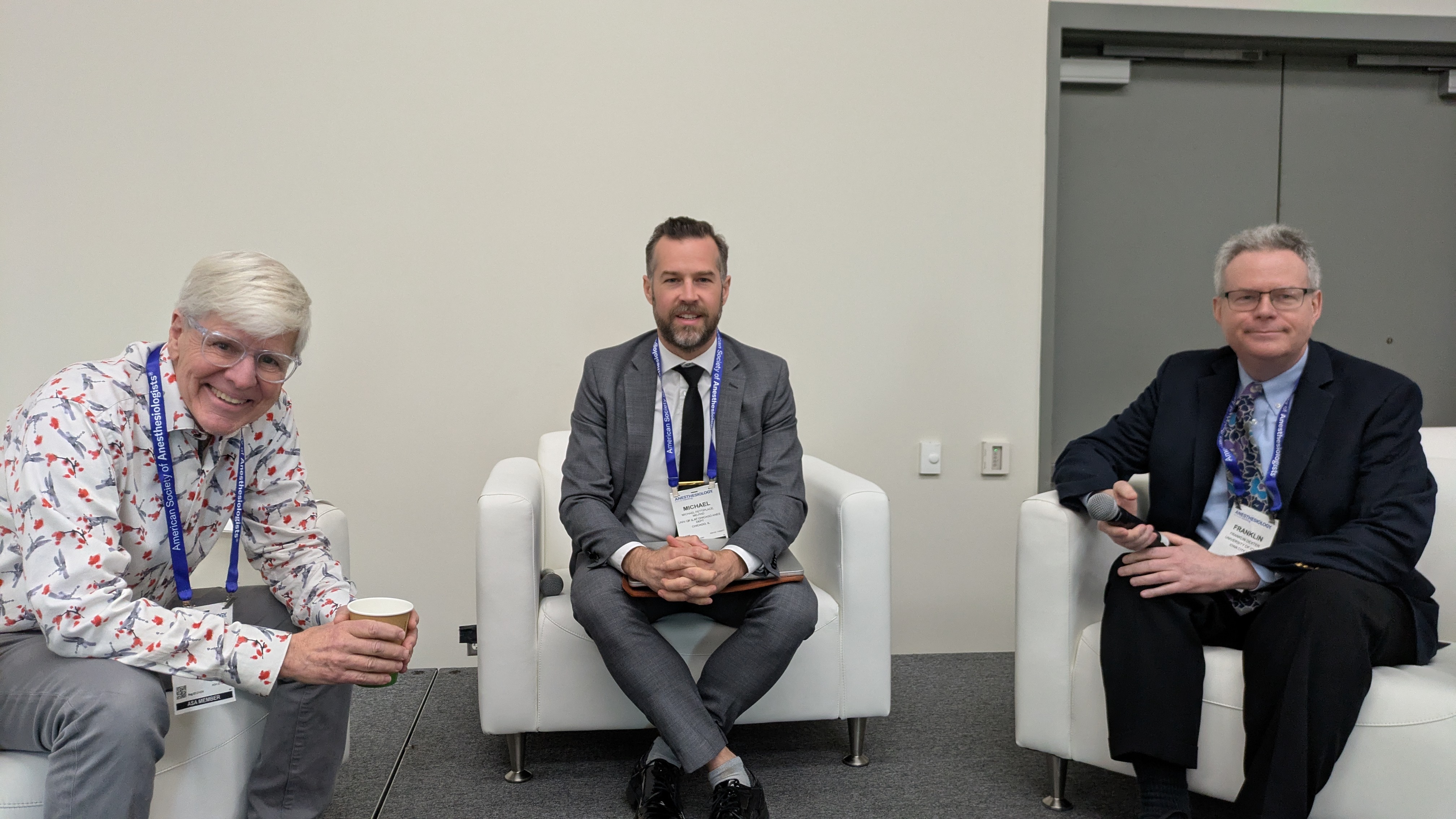 Three professionals wearing conference badges seated in white chairs during a panel discussion or networking event