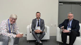 Three professionals wearing conference badges seated in white chairs during a panel discussion or networking event