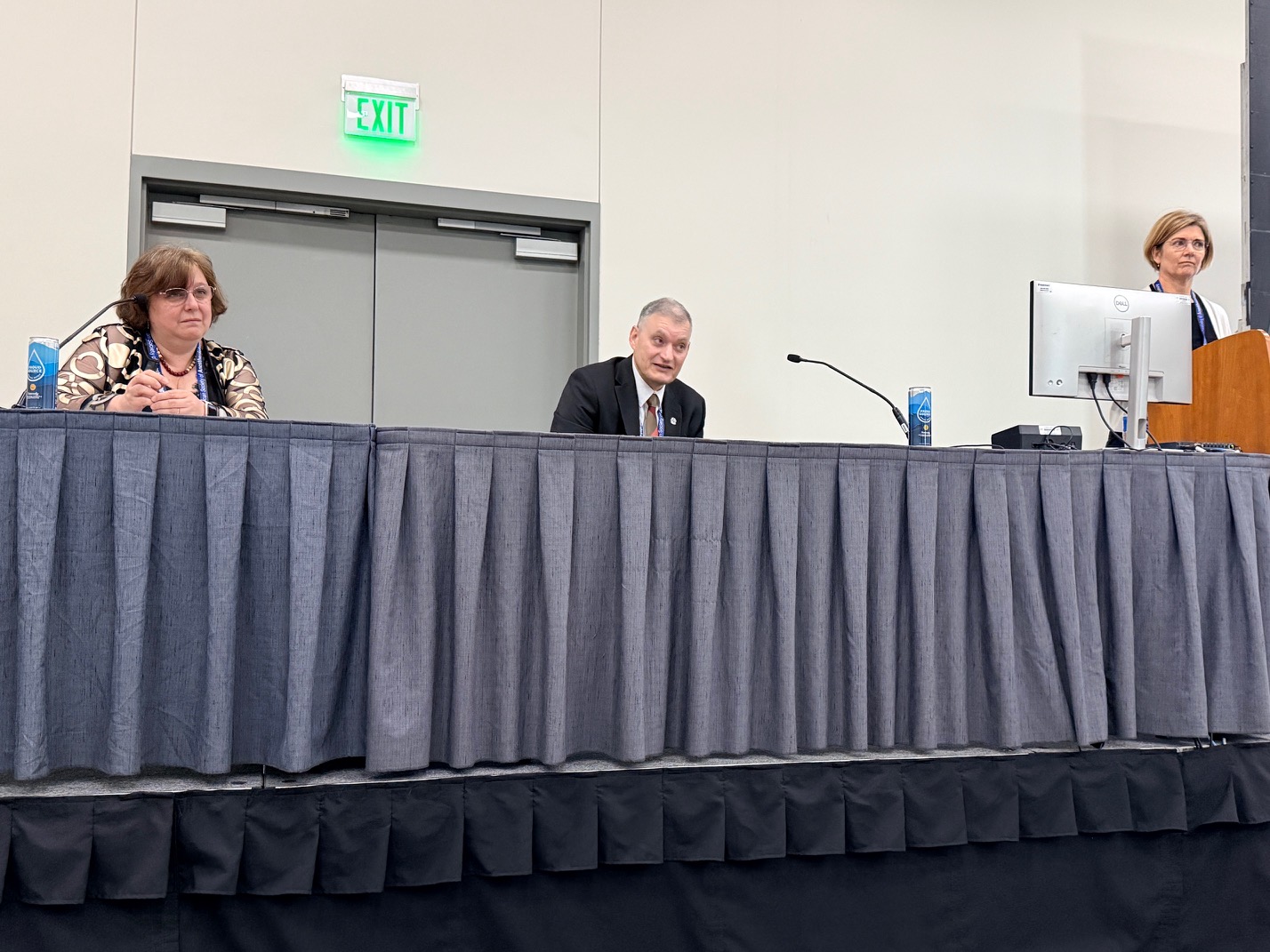 Three panelists seated at a draped table during a professional conference or panel discussion event.