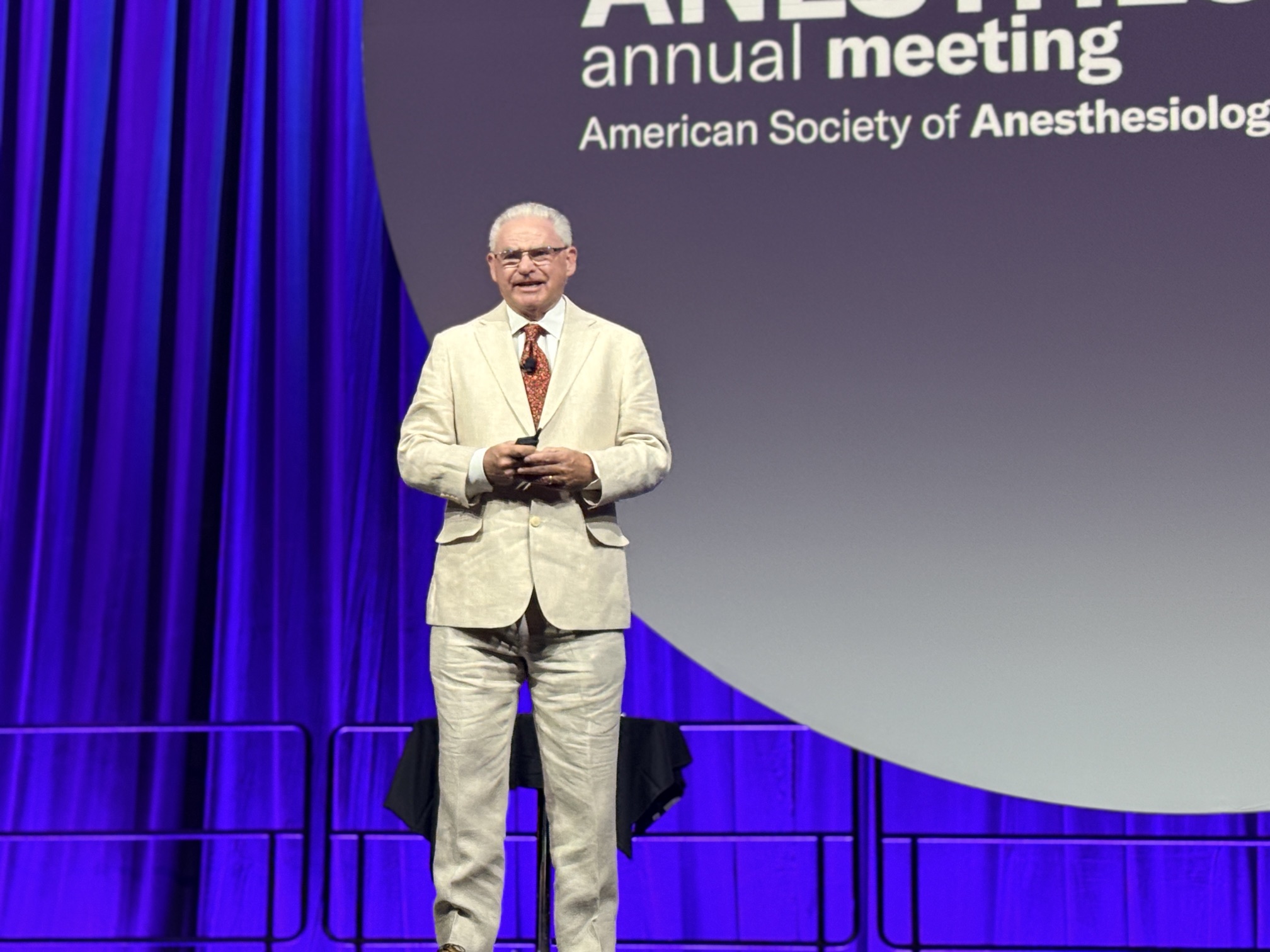 Presenter in cream suit speaking at podium during American Society of Anesthesiologists annual meeting with purple curtains and blue stage backdrop
