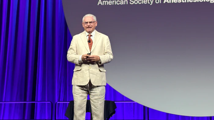 Presenter in cream suit speaking at podium during American Society of Anesthesiologists annual meeting with purple curtains and blue stage backdrop