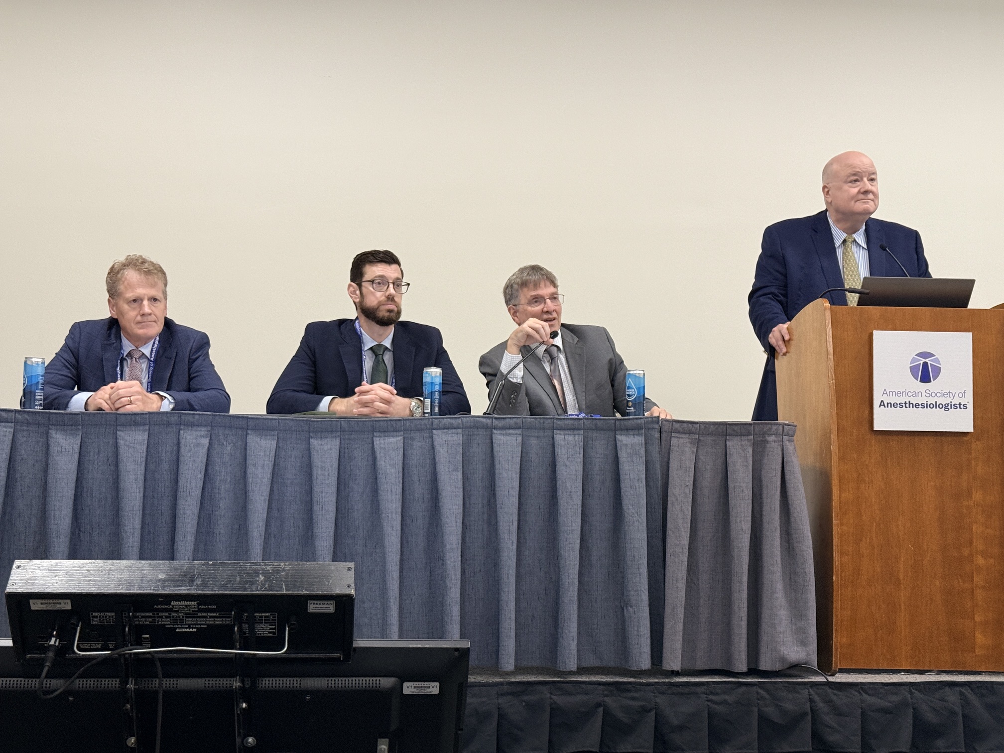 Four professionals at a panel discussion table with one speaker at podium displaying Anesthesiologists organization logo